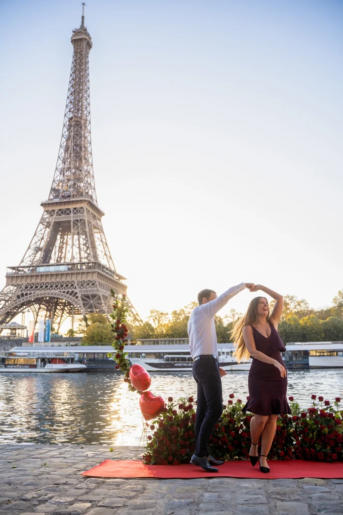 Public marriage proposal on the Seine quay across from the Eiffel Tower at sunrise, couple dancing beside red roses and heart balloons with tour boats behind them