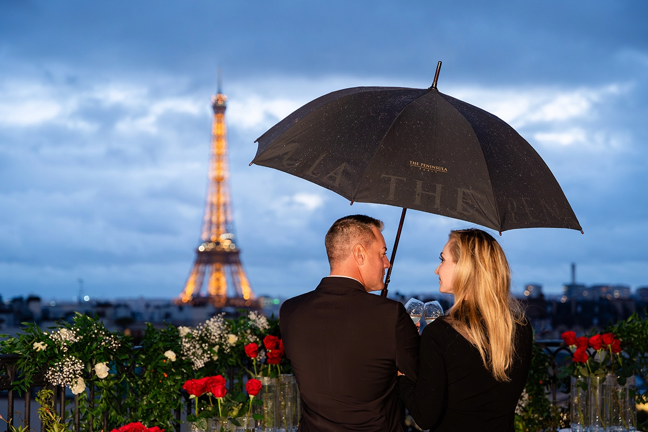 Couple sheltering under an umbrella on The Peninsula Paris rooftop terrace during rain, with the illuminated Eiffel Tower in the distance