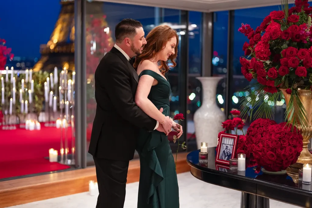 Man embracing his partner from behind as she holds a red rose, indoors with a red carpet, candles and the Eiffel Tower visible through the window