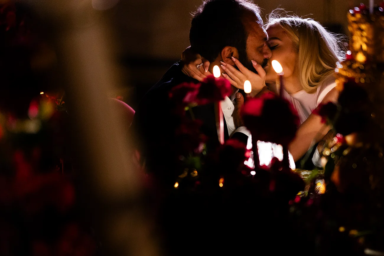 A newly engaged couple kisses at a candlelit private dinner surrounded by red roses and gold décor.