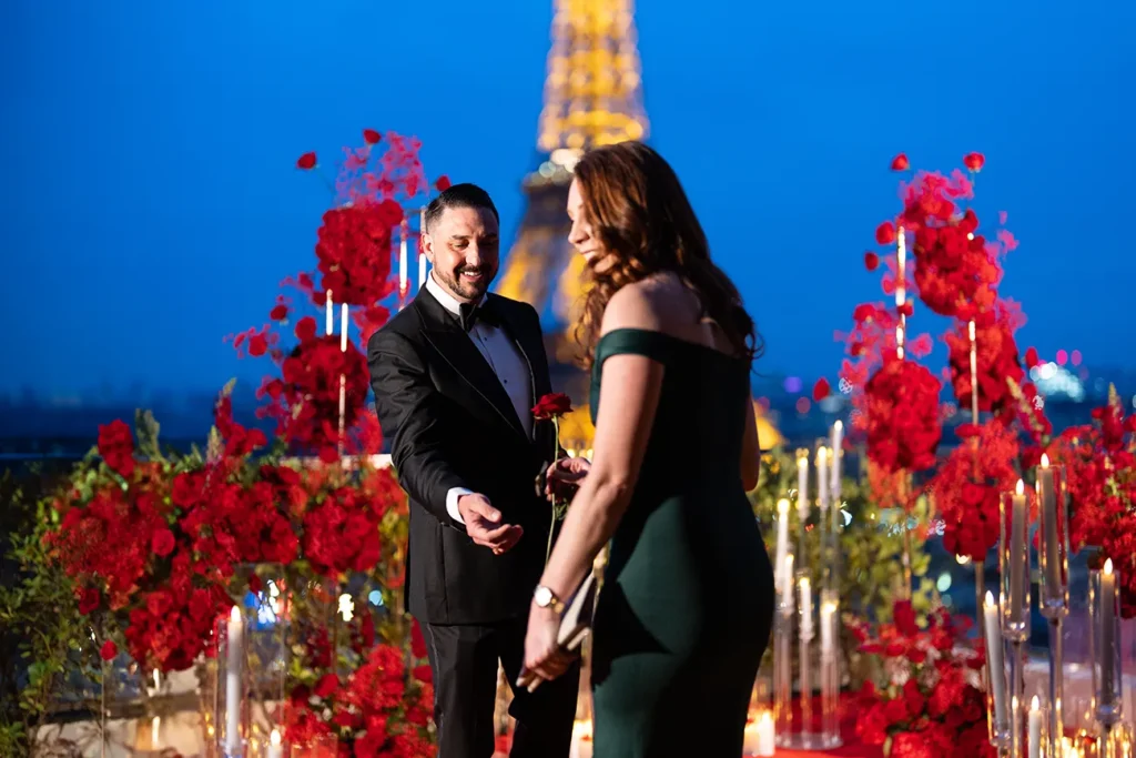 Engaged man presents a red rose to his partner during a rooftop proposal with the Eiffel Tower and red rose arrangements at La Suite Shangri La in Paris