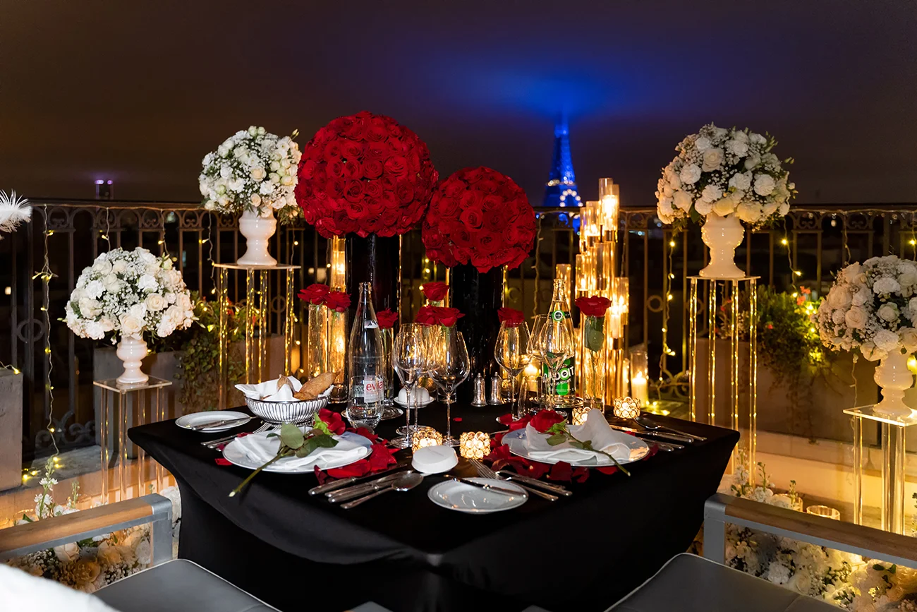 Private rooftop terrace at The Peninsula Paris set for a proposal dinner, with a black-linen table, red rose centerpieces, candlelight, and the illuminated Eiffel Tower in the distance