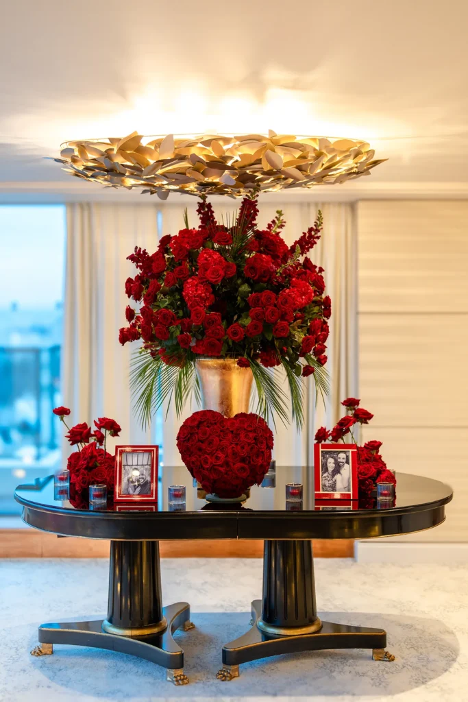 Tall red rose arrangement in a gold vase on a round entryway table with a rose heart, candles and framed couple photos below