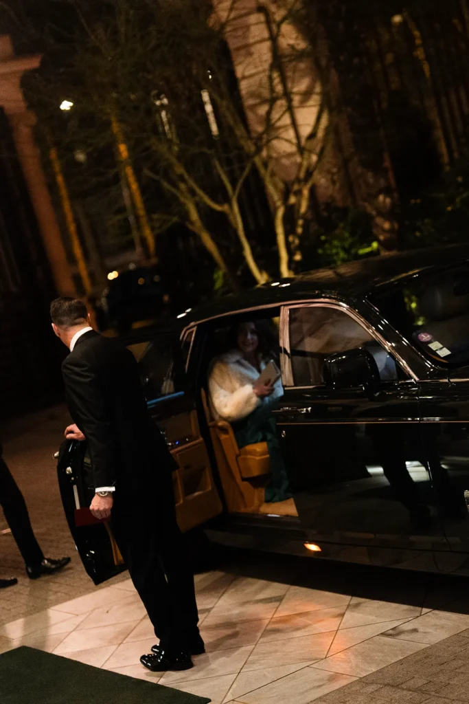 Woman seated inside a black Rolls Royce with the door held open by a staff member outside Shangri La Paris at night