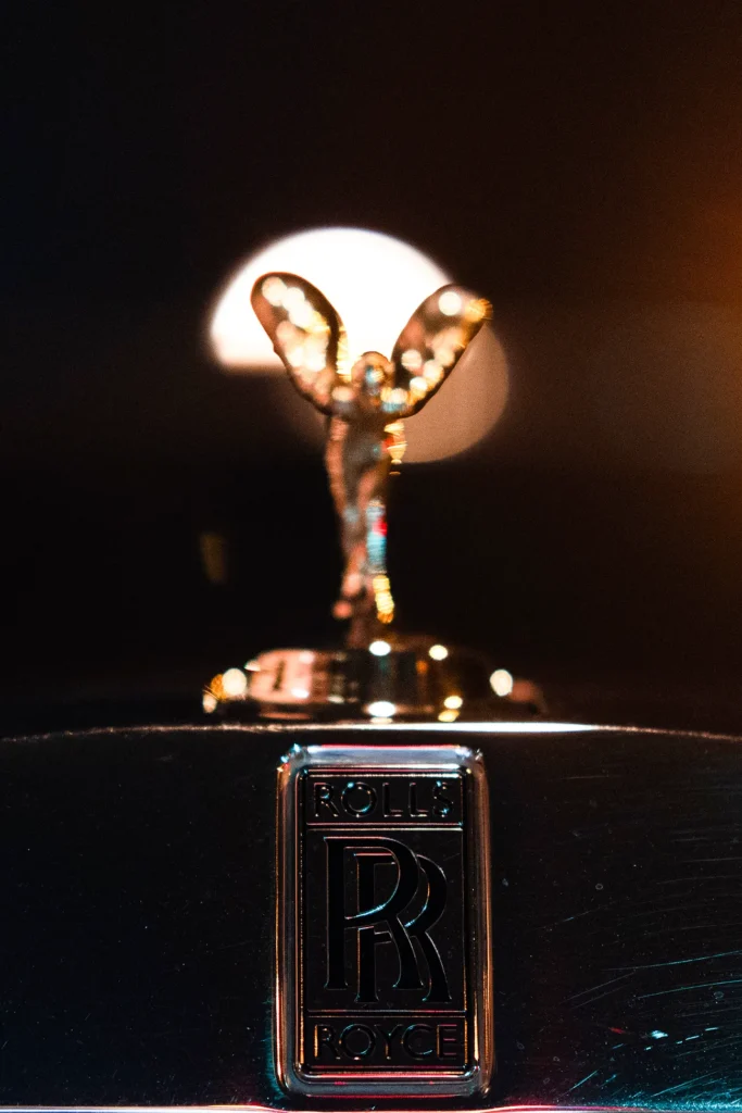 Close up of a Rolls Royce Spirit of Ecstasy hood ornament and grille badge lit by warm light at night