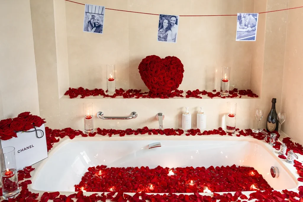 Hotel bathtub filled with red rose petals and floating candles, a red rose heart, Chanel gift bag, champagne and a string of couple photos above