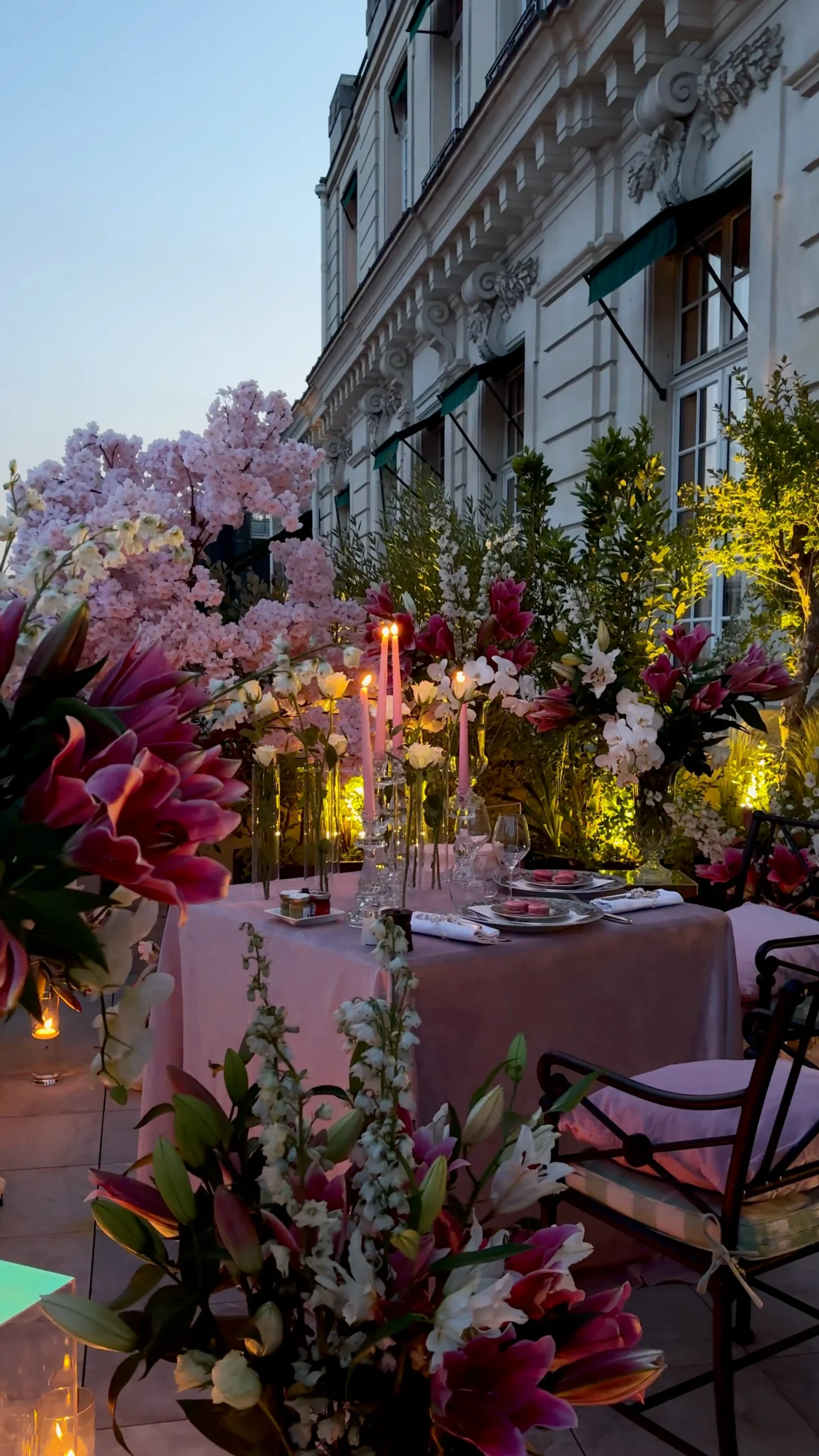 Private dusk dinner table at Shangri-La Paris set with pink candles, macarons, and champagne glasses, surrounded by dense arrangements of magenta lilies and pink cherry blossom against the hotel façade