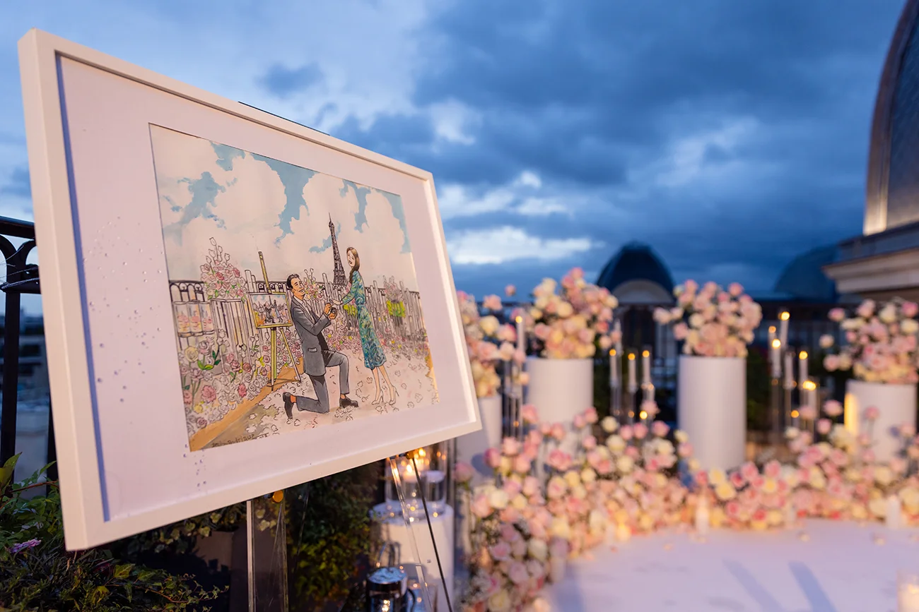 Custom illustrated portrait of a proposal on The Peninsula Paris rooftop terrace, displayed on an easel beside fresh rose arrangements with the Eiffel Tower in the background at dusk