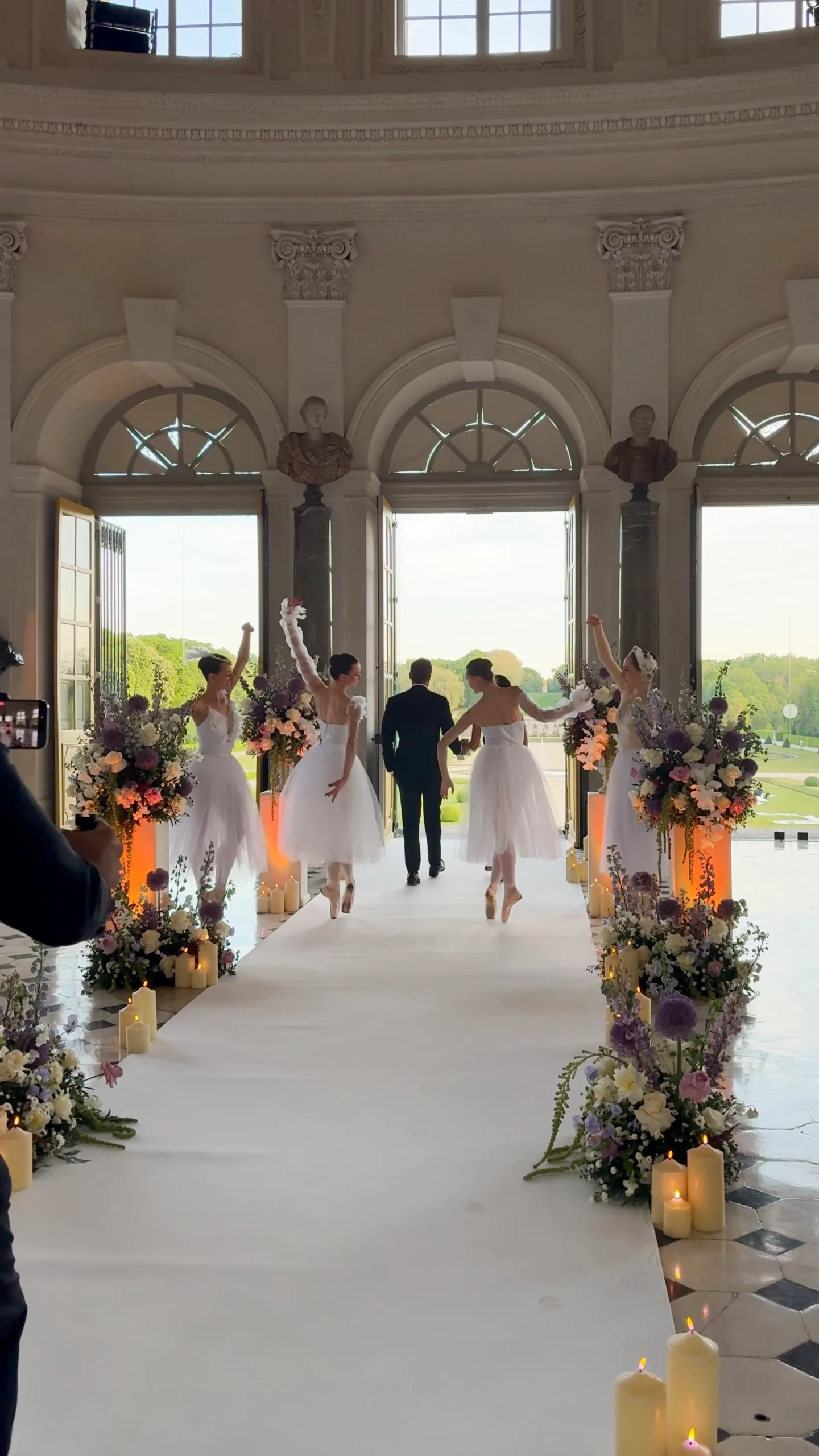 Ballerinas in white tutus performing along a candlelit aisle inside the grand hall of Château de Vaux-le-Vicomte as a man in a tuxedo walks toward the open château doors overlooking the gardens