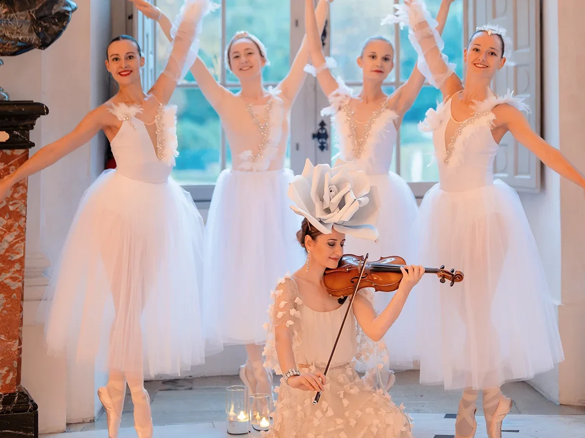Solo violinist in floral headpiece performs with four ballerinas in front of tall château windows